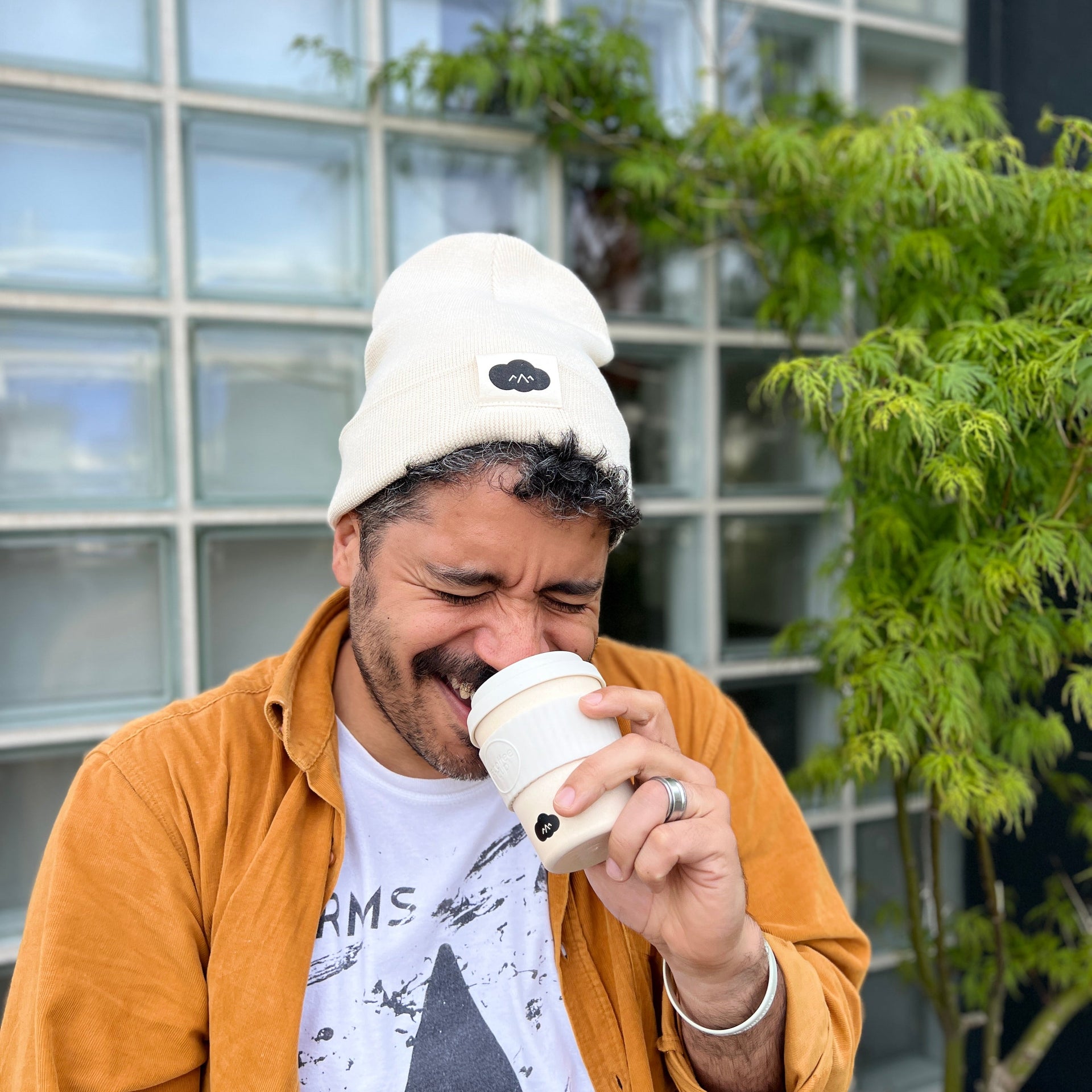 Man drinking from a coffee cup outdoors with a building and plants in the background