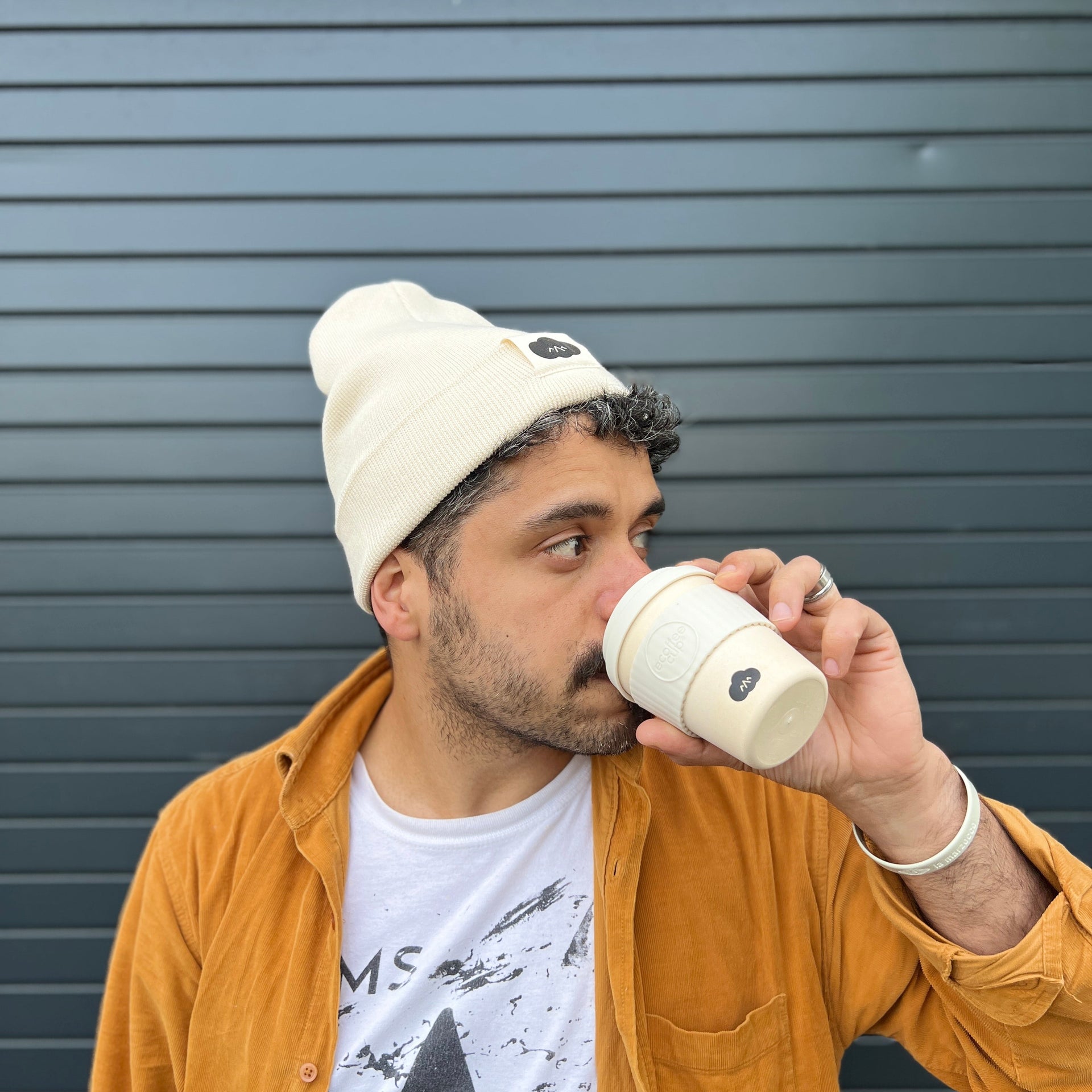 Man drinking from a coffee cup against a gray background in a beige beanie