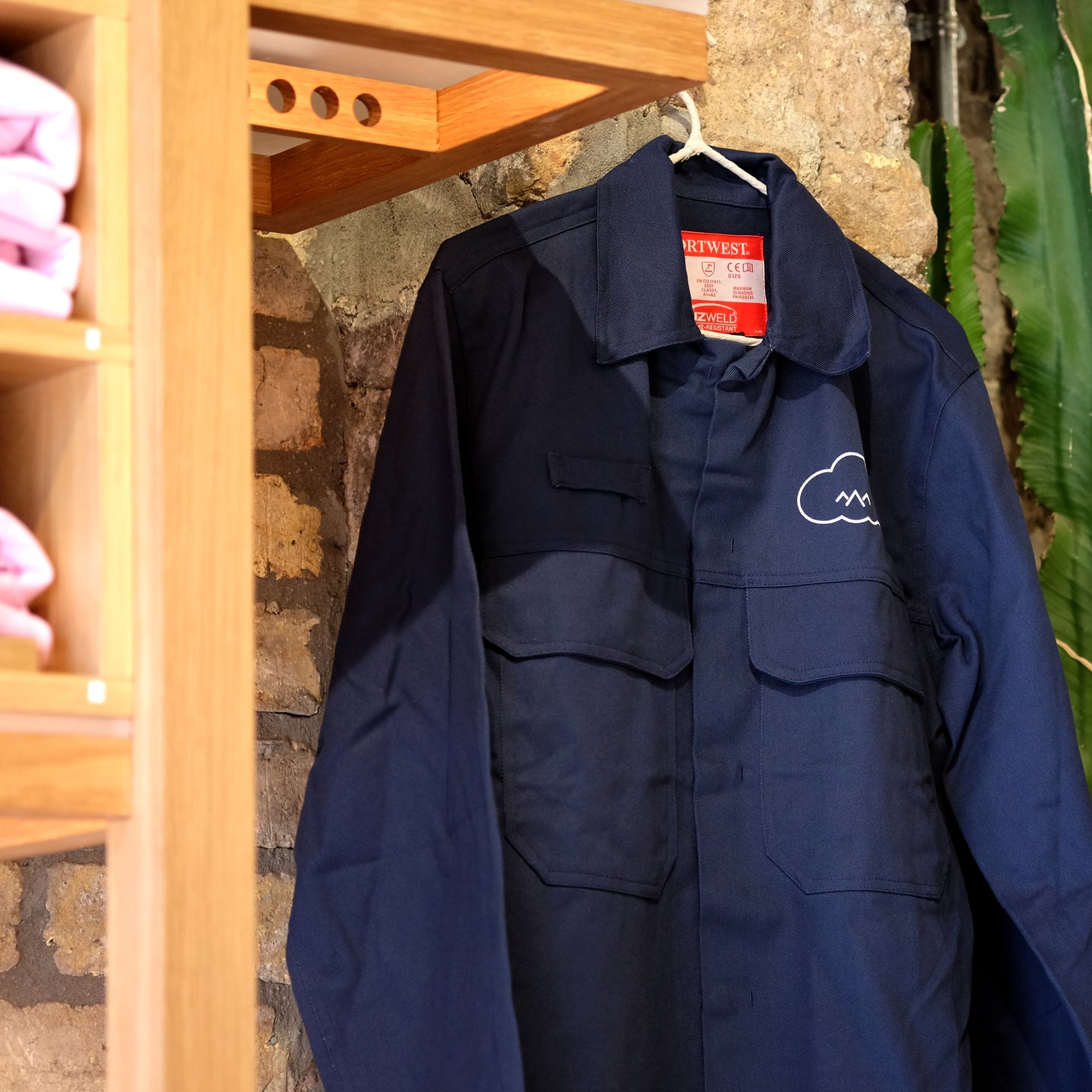 Unisex Cloud Picker Coffee roaster jacket hanging on a cafe wall, showcasing durable merchandise for coffee professionals.